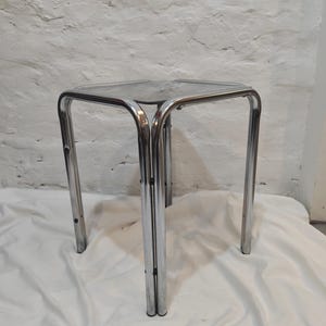 Könnte beinhalten: Ein Beistelltisch aus verchromtem Metall mit quadratischer Glasplatte. Die Beine sind gebogen und röhrenförmig, an den Ecken verbunden. Der Tisch steht auf einer weißen Stoffoberfläche vor einer weißen Backsteinmauer.