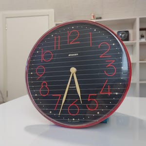 May include: A red and black analog clock with gold hands and a white face. The clock has black lines and red numbers around the edge. The clock says "QUARTZ" and "WEST GERMANY" on the face.