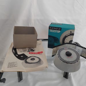 Può includere: Un timer vintage Kienzle Telestop con un quadrante bianco e nero e un quadrante bianco. Il timer è su una superficie bianca con una scatola marrone e una scatola blu e bianca con il testo "Kienzle Telestop" su di essa.