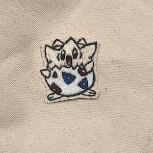 May include: Embroidered patch featuring a Togepi character from the Pokémon series. The patch has a white, blue, red, and black color scheme. The patch is sewn onto a beige fabric background.