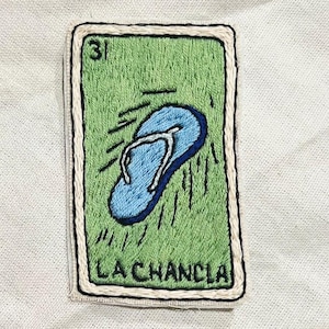 May include: Embroidered patch featuring a blue flip-flop on a green background, with the number 31 at the top. The text "LA CHANCLA" is at the bottom, all within a rectangular frame.