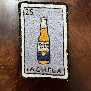 May include: Embroidered Loteria card with a Corona Extra beer bottle on a gray background. The card is numbered 25 and the text "LA CHELA" is at the bottom.