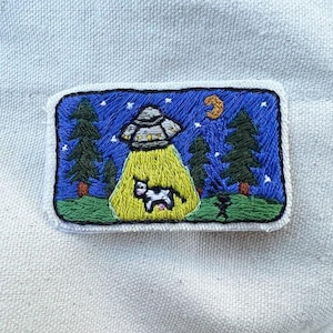 May include: Embroidered patch featuring a UFO beaming up a cow. The patch has a blue night sky with stars, green trees, and a crescent moon. A small figure stands on the ground. The patch is rectangular with a white border.