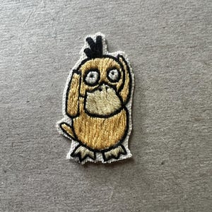 May include: Embroidered patch featuring a cartoon duck-like character with a yellow body, black outlines, and large, wide eyes. The patch has a beige border and is likely designed for application on clothing or accessories.