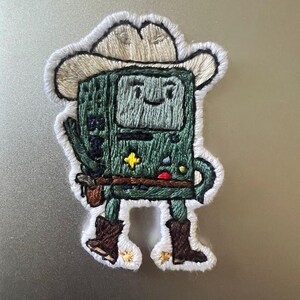 May include: Embroidered patch of a green cartoon character wearing a cowboy hat and boots. The character has a smiling face and is holding a gun in its right hand.