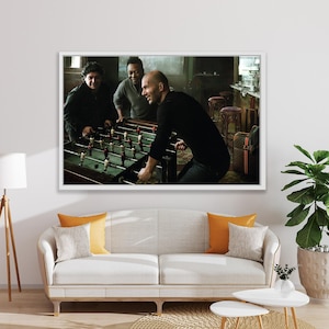 Könnte beinhalten: Ein gerahmter Druck zeigt drei Männer beim Tischfußball. Der Tischfußballtisch hat rote und weiße Spielerfiguren. Die Männer sind lässig gekleidet. Der Hintergrund umfasst eine Bar und Hocker. Der Druck hängt über einem weißen Sofa mit orangefarbenen Kissen.