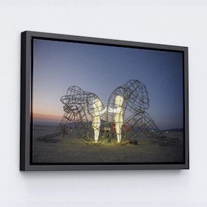 Puede incluir: Impresión enmarcada que presenta una escultura de dos figuras de alambre, iluminadas desde dentro. Las figuras están sentadas, una frente a la otra, contra un cielo crepuscular. La obra de arte está sobre un terreno arenoso, con un marco negro.