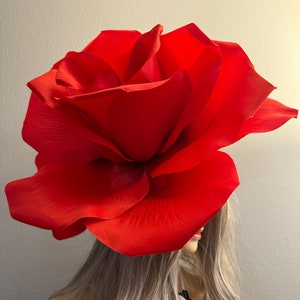 Op de afbeelding: Een grote, rode rozenhoed van stof. De roos wordt op het hoofd van een persoon gedragen.