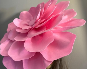 Könnte beinhalten: Ein großer rosa Hut aus Filz in Blumenform. Der Hut besteht aus vielen Schichten von Blütenblättern und ist zum Tragen auf dem Kopf gedacht.