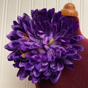 May include: A large, purple silk flower with a layered, ruffled design. The flower has a slightly darker purple hue in the center and is accented with small, gold-colored accents.