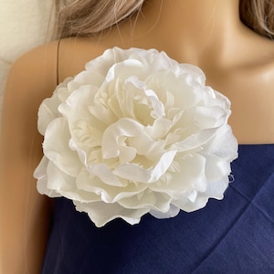 May include: A large, white, silk flower brooch. The flower has a layered, ruffled appearance and is reminiscent of a peony.