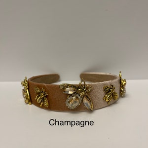 May include: A champagne-colored headband with a velvet texture and gold bee embellishments. The headband has a white satin stripe and features a large crystal bee in the center.