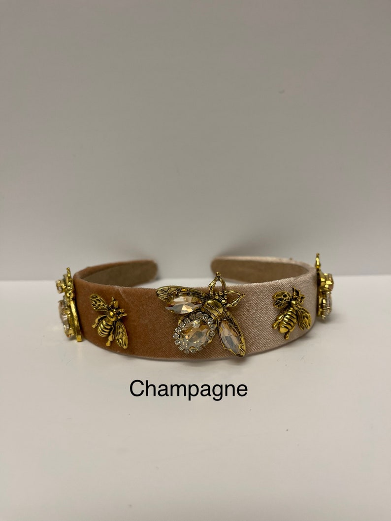 May include: A champagne-colored headband with a velvet texture and gold bee embellishments. The headband has a white satin stripe and features a large crystal bee in the center.