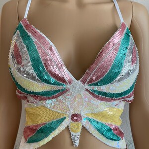 Puede incluir: Un top corto blanco con un dise&ntilde;o de mariposa hecho de lentejuelas. La mariposa es multicolor con lentejuelas rosas, verdes, amarillas y blancas.