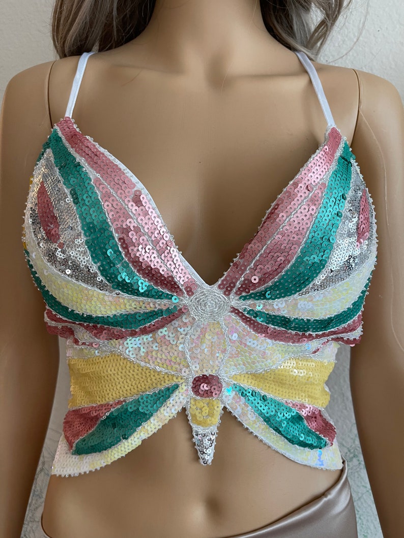 Puede incluir: Un top corto blanco con un dise&ntilde;o de mariposa hecho de lentejuelas. La mariposa es multicolor con lentejuelas rosas, verdes, amarillas y blancas.