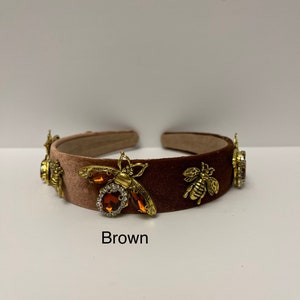 May include: A brown velvet headband with gold bee embellishments and brown rhinestones. The headband is decorated with three bees and rhinestones.