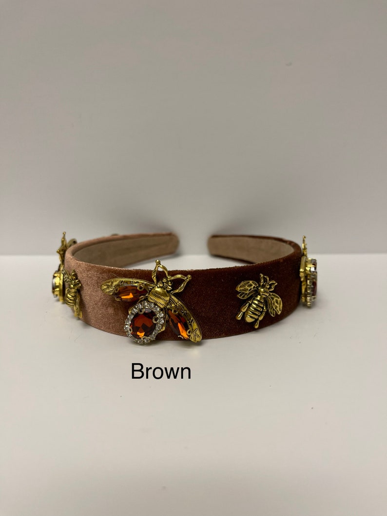 May include: A brown velvet headband with gold bee embellishments and brown rhinestones. The headband is decorated with three bees and rhinestones.
