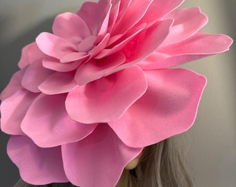 Könnte beinhalten: Ein großer, rosa Blütenkopfschmuck. Die Blütenblätter sind ein weiches, einfarbiges Rosa, und die Blume wird auf dem Kopf getragen. Der Kopfschmuck ist ein Statement-Stück, perfekt für einen besonderen Anlass oder ein Kostüm.