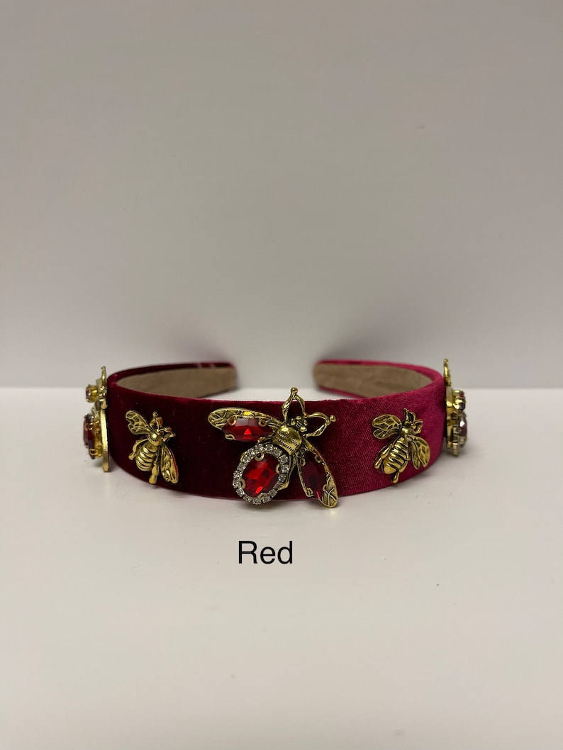 May include: A burgundy velvet headband with gold and red bee embellishments. The headband is decorated with rhinestones and has a red jewel in the center.