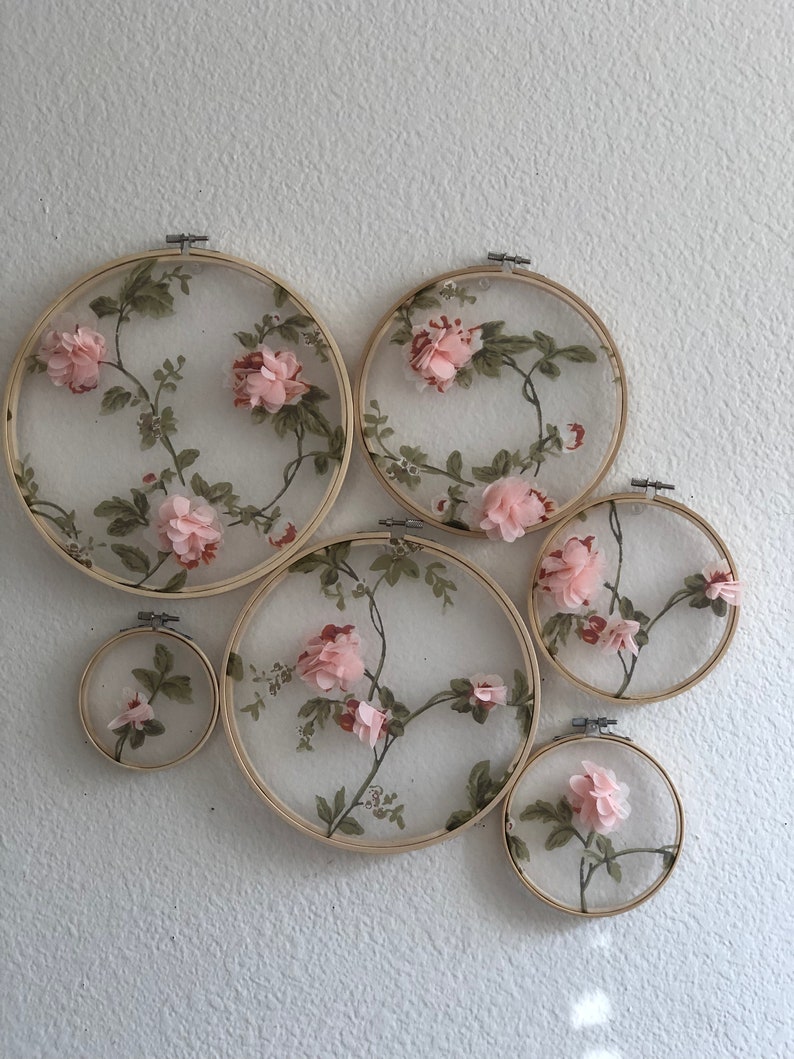 May include: A set of six embroidery hoops with floral designs. The hoops are made of wood and have a natural finish. The floral designs are made of pink and green fabric and are arranged in a circular pattern.