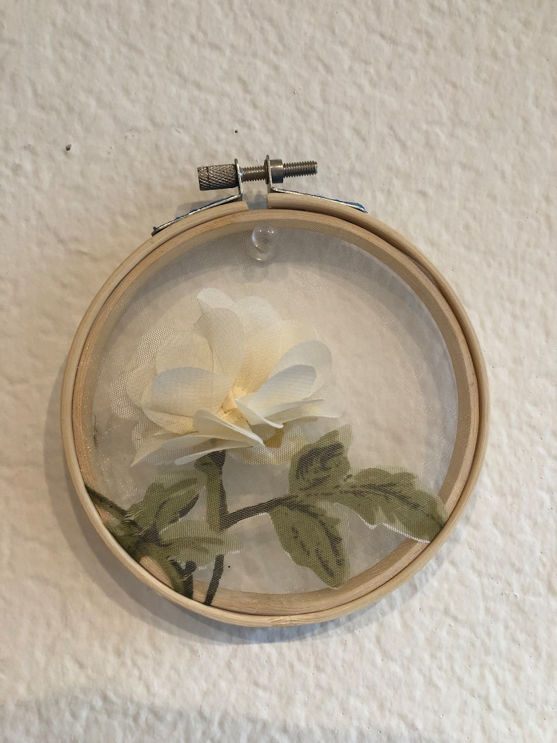 May include: A white fabric flower with green leaves is displayed in a wooden embroidery hoop.