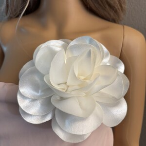 May include: A white satin flower brooch with multiple layers of petals. The flower is attached to a pink fabric background.