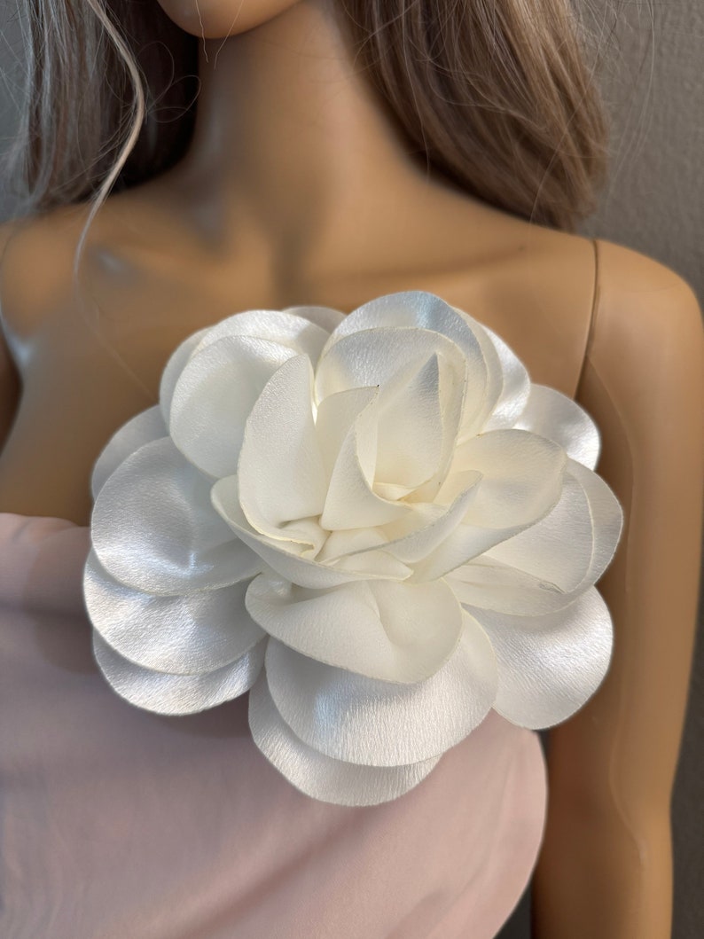 May include: A white satin flower brooch with multiple layers of petals. The flower is attached to a pink fabric background.