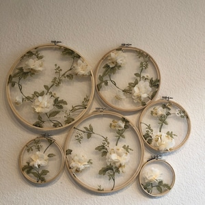 May include: Six round embroidery hoops with floral fabric designs. The hoops are various sizes and are arranged in a cluster on a white wall.