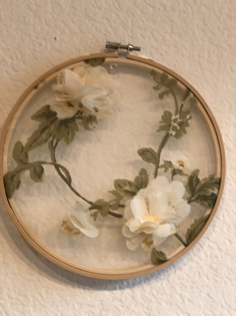 May include: A white floral embroidery design in a wooden embroidery hoop. The flowers are white and the leaves are green.