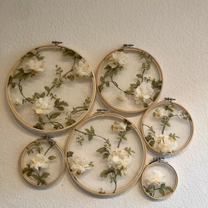 May include: Six round embroidery hoops with white floral fabric stretched over them. The hoops are arranged in a staggered pattern on a white wall.