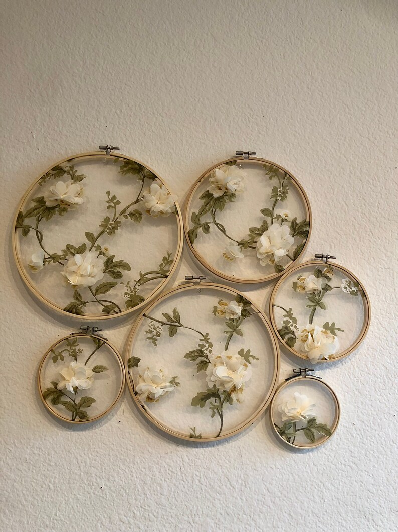 May include: Six round embroidery hoops with white floral fabric stretched over them. The hoops are arranged in a staggered pattern on a white wall.