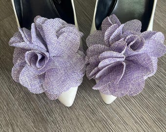 May include: A pair of white high heels with pointed toes, each adorned with a large, lavender fabric flower. The flowers have a textured, layered petal design. The shoes are set against a gray wooden surface.