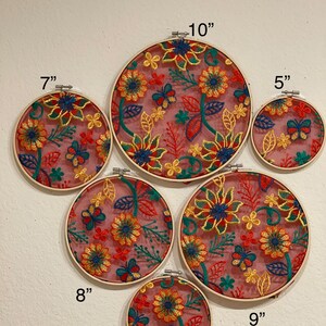 May include: Set of six embroidery hoops with floral designs in shades of red, yellow, green, and blue on a pink background. The hoops are 5, 6, 7, 8, 9, and 10 inches in diameter.