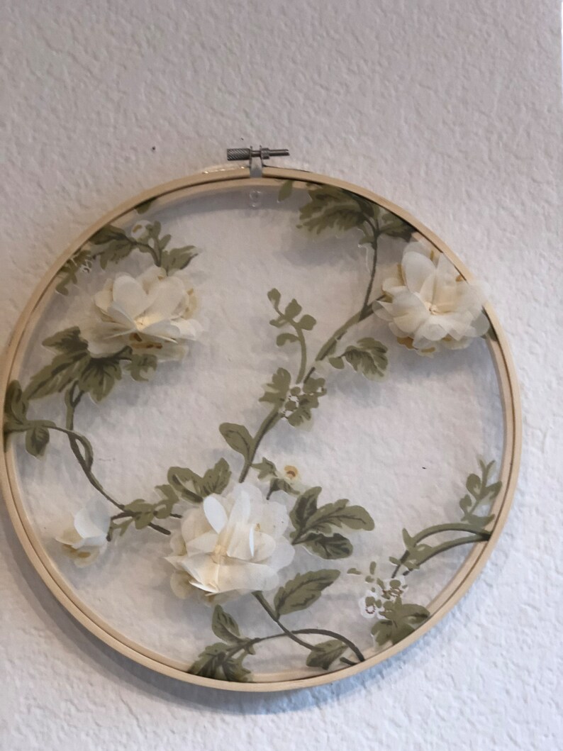 May include: A white floral embroidery design in a wooden embroidery hoop. The design features white flowers and green leaves on a white background.
