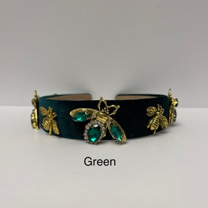May include: A dark green velvet headband with gold bee embellishments and green gem accents. The headband has the word "Green" written below it.