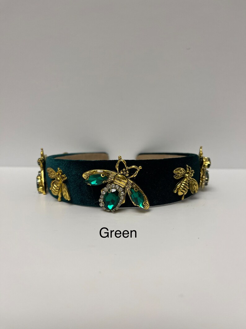 May include: A dark green velvet headband with gold bee embellishments and green gem accents. The headband has the word "Green" written below it.