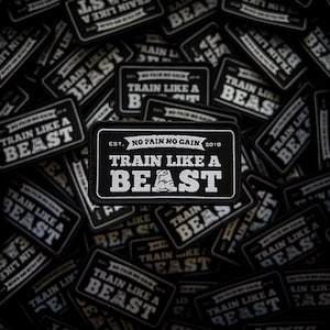 Peut inclure: Écussons brodés noirs et blancs avec le texte "TRAIN LIKE A BEAST" et "NO PAIN NO GAIN 2018". Les écussons présentent un graphique d'haltérophile et sont de forme rectangulaire. Thème fitness et entraînement.
