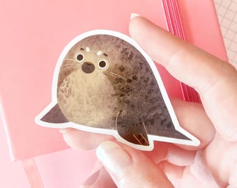 Puede incluir: Una pegatina troquelada que muestra una foca redonda de color marrón con manchas, vientre de color tostado claro, ojos grandes y oscuros y bigotes largos. La foca tiene una textura ilustrada suave con un borde blanco y está sentada.