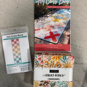 May include: An assortment of sewing supplies, including a table runner pattern, fabric squares, and paper pieces. The table runner pattern is titled "Hot Cross Buns" and features a red cross design. The fabric squares are labeled "Sunday Brunch".