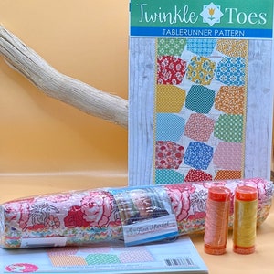 May include: A table runner quilting pattern called "Twinkle Toes" with a colorful patchwork design. The pattern is accompanied by fabric scraps, thread spools, and a book.