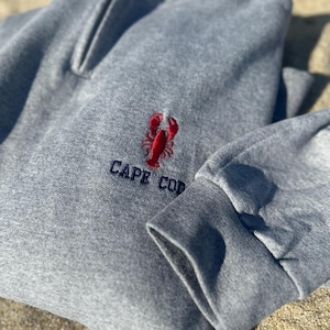 May include: A grey sweatshirt featuring a red embroidered lobster and the words "CAPE COD" in navy blue. The sweatshirt has a ribbed cuff and a drawstring neckline, suitable for casual wear.