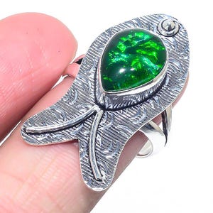 May include: A silver ring with a large, pear-shaped, vivid green gemstone. The ring has an intricate, textured design with a floral pattern. The gemstone is set in a bezel setting, and the ring's band is a simple, polished silver.