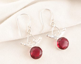 May include: A pair of silver earrings with a hook design. Each earring features a round, deep red gemstone. The earrings have a decorative, branch-like element above the gemstone, adding a unique touch to the jewelry.