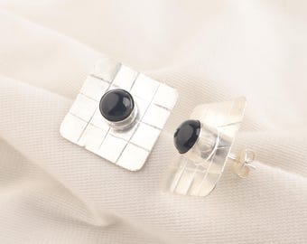 May include: A pair of silver square earrings with black round gemstones in the center.
