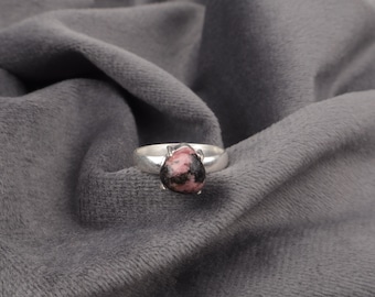 Pode incluir: Um anel de prata com uma gema rosa e preta. O anel está sobre um fundo de tecido cinza.