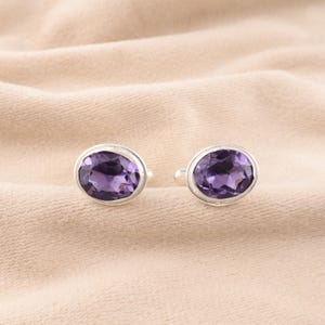May include: A pair of silver cufflinks, each featuring a large, oval-cut, deep purple amethyst gemstone. The gemstones are set in a polished silver bezel, creating a classic and elegant design. The cufflinks are displayed on a neutral, textured background.