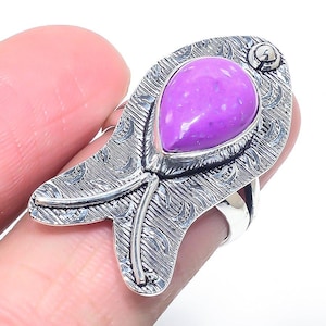 May include: A silver-toned ring featuring a large, teardrop-shaped, speckled purple stone. The ring's setting has an ornate, textured design with a floral motif. The ring is held by a person's finger.