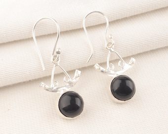May include: A pair of silver earrings featuring a black, round gemstone. The earrings have a decorative silver design above the gemstone and a simple hook for the ear. The background is a white, textured fabric.