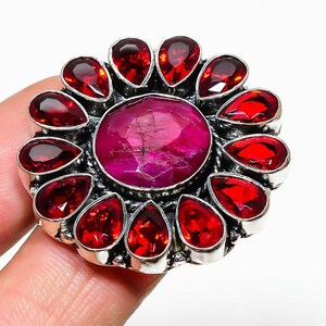May include: A silver ring featuring a large, oval, pink-red gemstone at the center, surrounded by a halo of smaller, teardrop-shaped, red gemstones. The ring has a detailed, ornate setting.