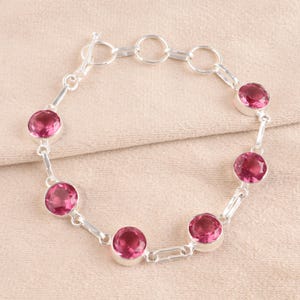 May include: A silver bracelet with a lobster clasp and five pink round gemstones. The bracelet is on a light pink fabric background.